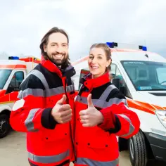 Uśmiechnięci ratownicy medyczni w jaskrawych kurtkach, z kciukami w górze, na tle karetki. Gotowi do akcji, zapewniają profesjonalne umed ratownictwo medyczne.