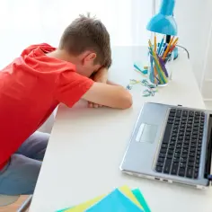 Chłopiec w czerwonej koszulce śpi na biurku obok laptopa i przyborów szkolnych.