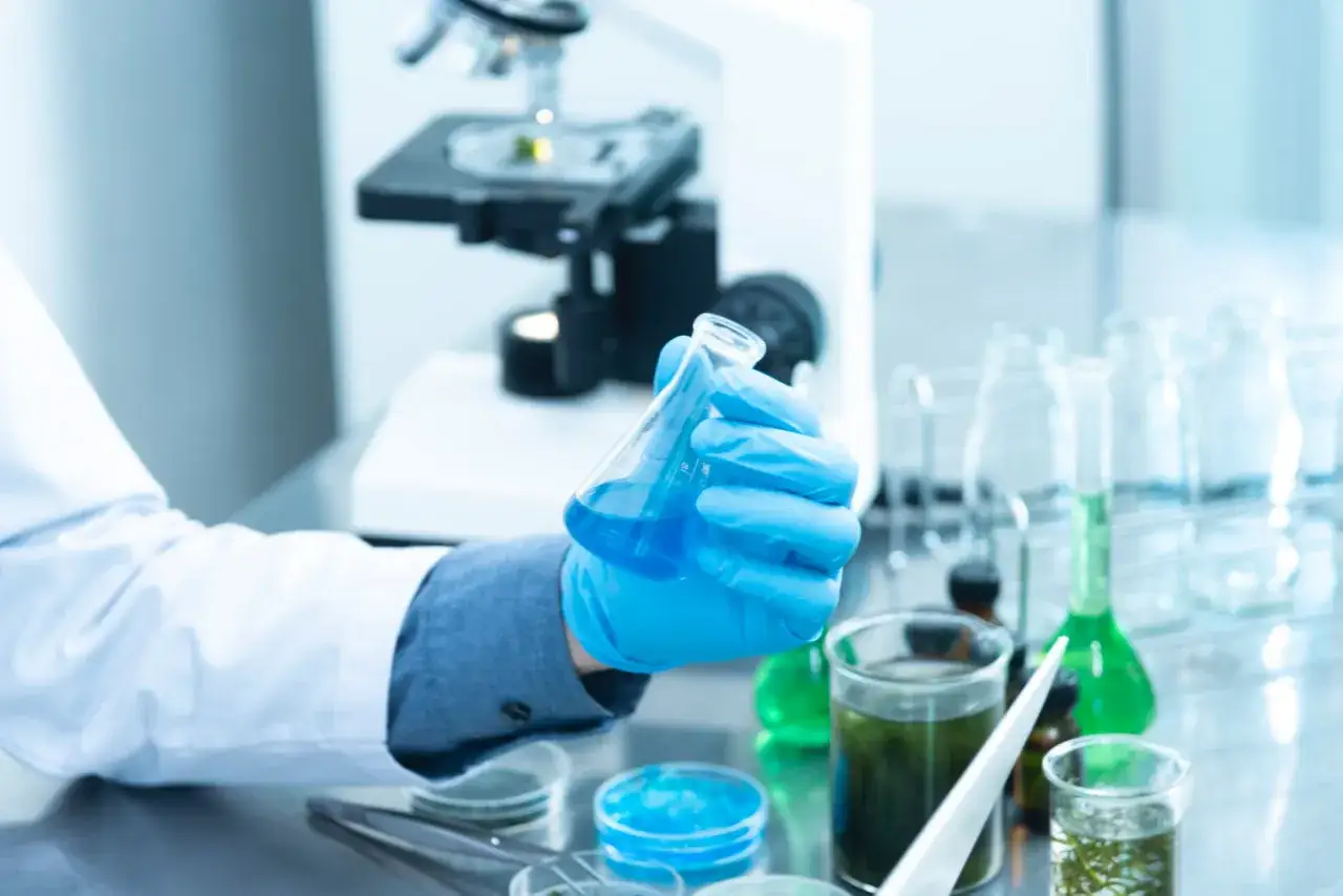Ręka w niebieskiej rękawiczce trzyma kolbę z niebieskim płynem. W tle mikroskop i inne naczynia laboratoryjne. To początek badań, co po chemii.